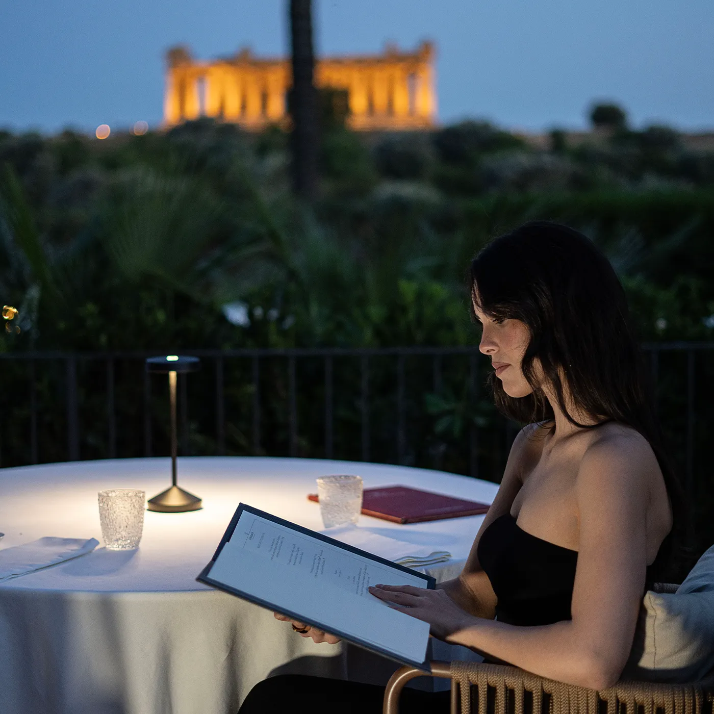 Cena romantica al tramonto a Villa Athena Resort, con donna che legge il menu e il Tempio della Concordia illuminato sullo sfondo.