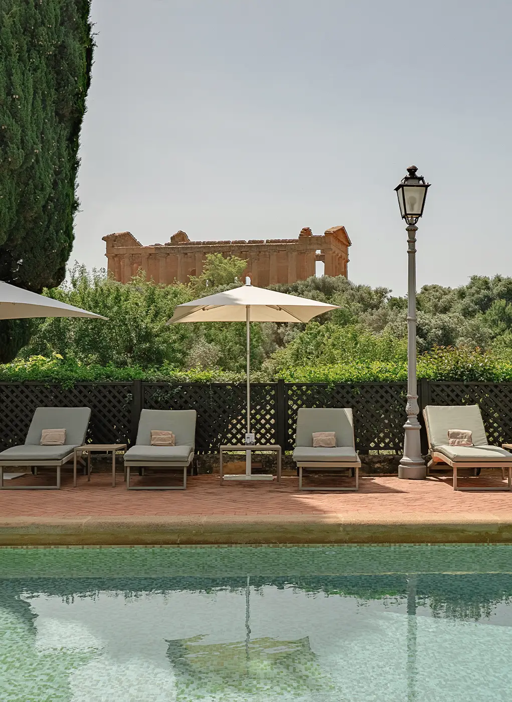 Area solarium a bordo piscina di Villa Athena Resort con lettini, ombrellone e il Tempio della Concordia sullo sfondo.