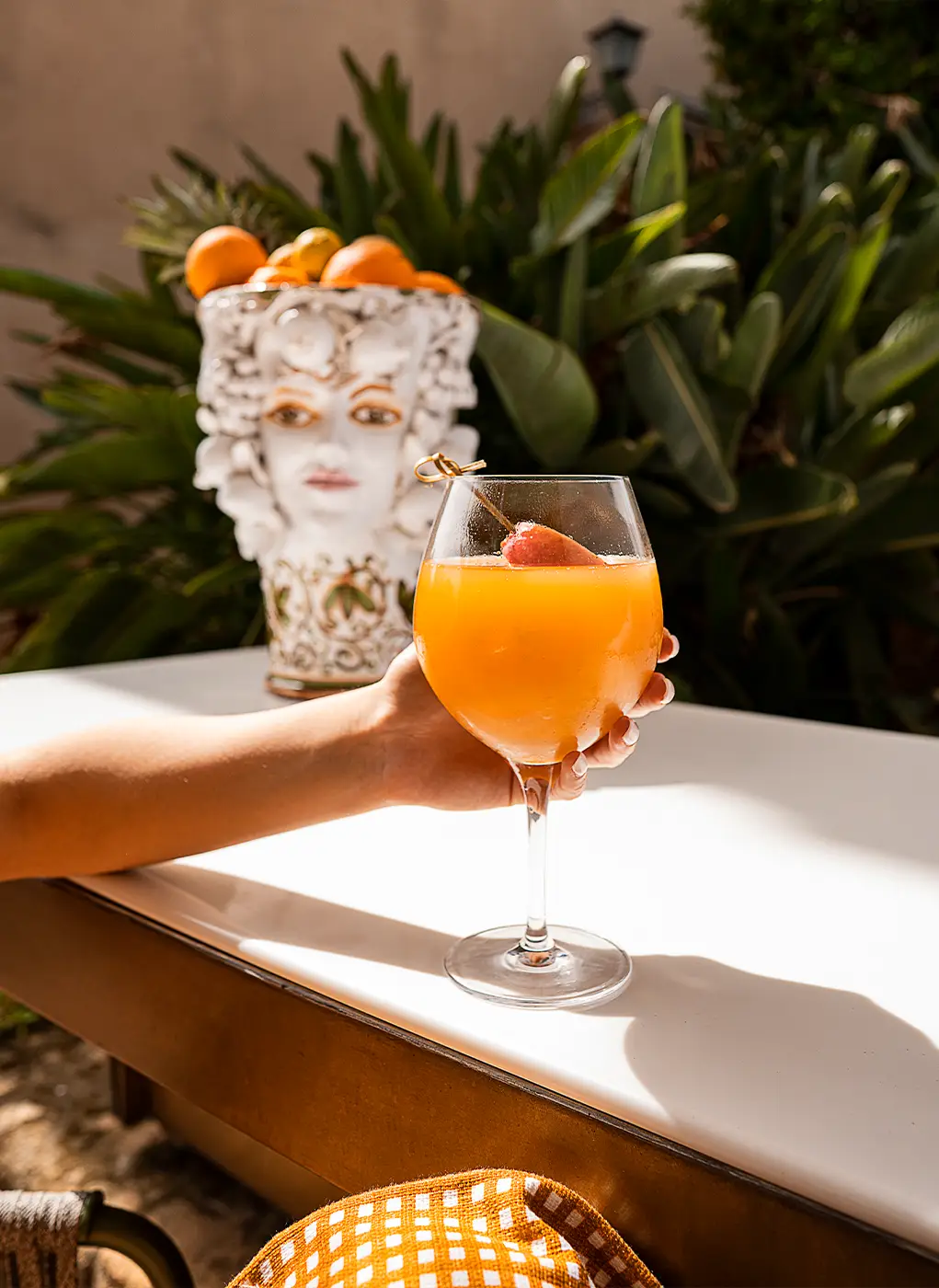 Mano che regge un cocktail all'arancia al Villa Athena Resort, con una testa di Moro siciliana piena d'agrumi sullo sfondo.